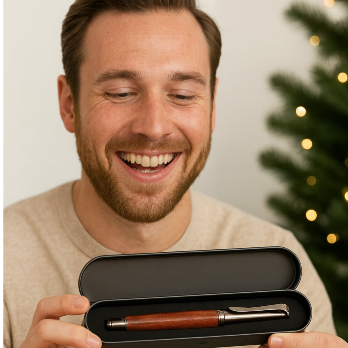 Homme souriant qui découvre un stylo en bois à finitions métal gun dans son écrin en métal noir, devant un sapin de Noël – Weastyle Gravure
