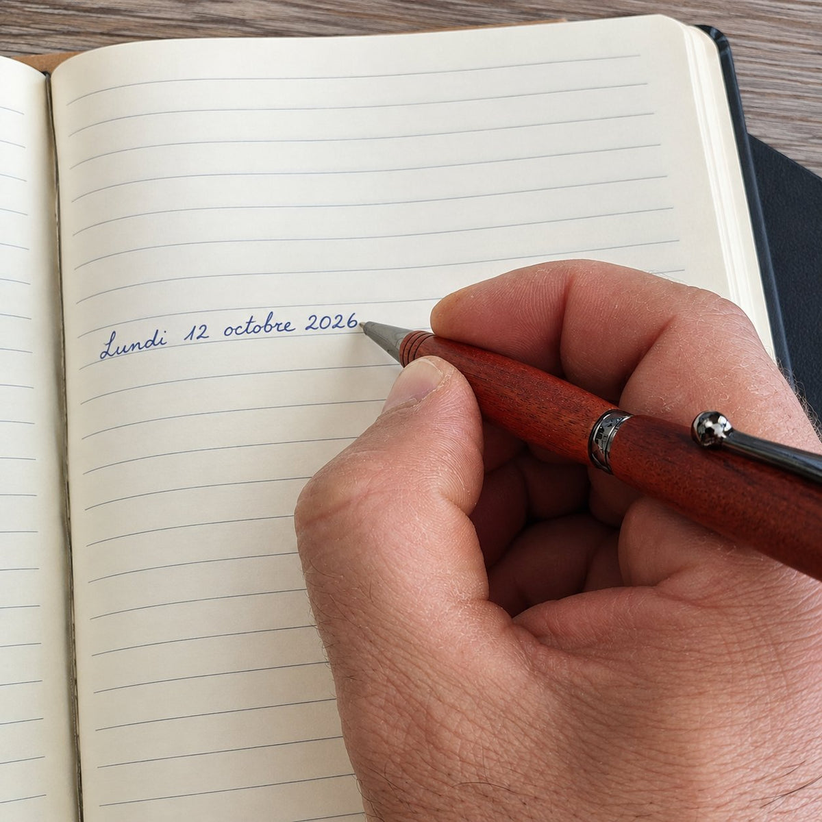 Main qui écrit à l'encre bleue dans un carnet ligné avec un stylo en bois rotatif à clip métal – Weastyle Gravure