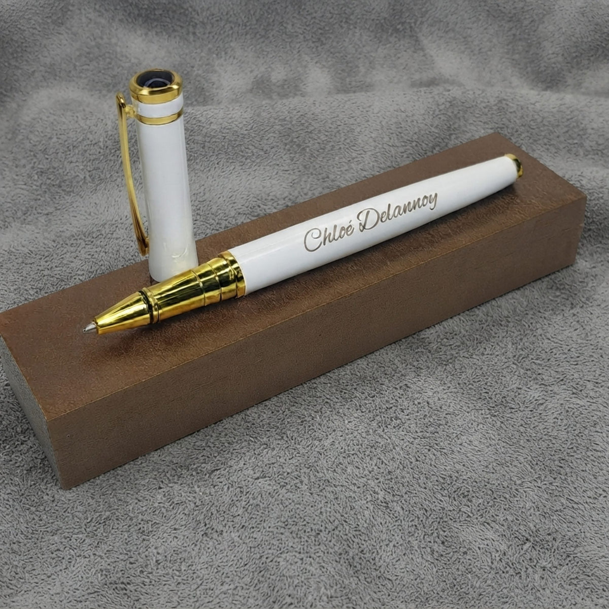 Stylo en métal blanc et doré gravé « Chloé Delannoy », capuchon à clip doré posé debout, sur un socle brun et un fond de feutre gris – Weastyle Gravure